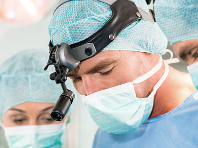 Ein Chirurg mit einer Kopfbeleuchtung, umgeben von weiteren medizinischen Fachleuten, alle tragen OP-Kleidung und Masken.